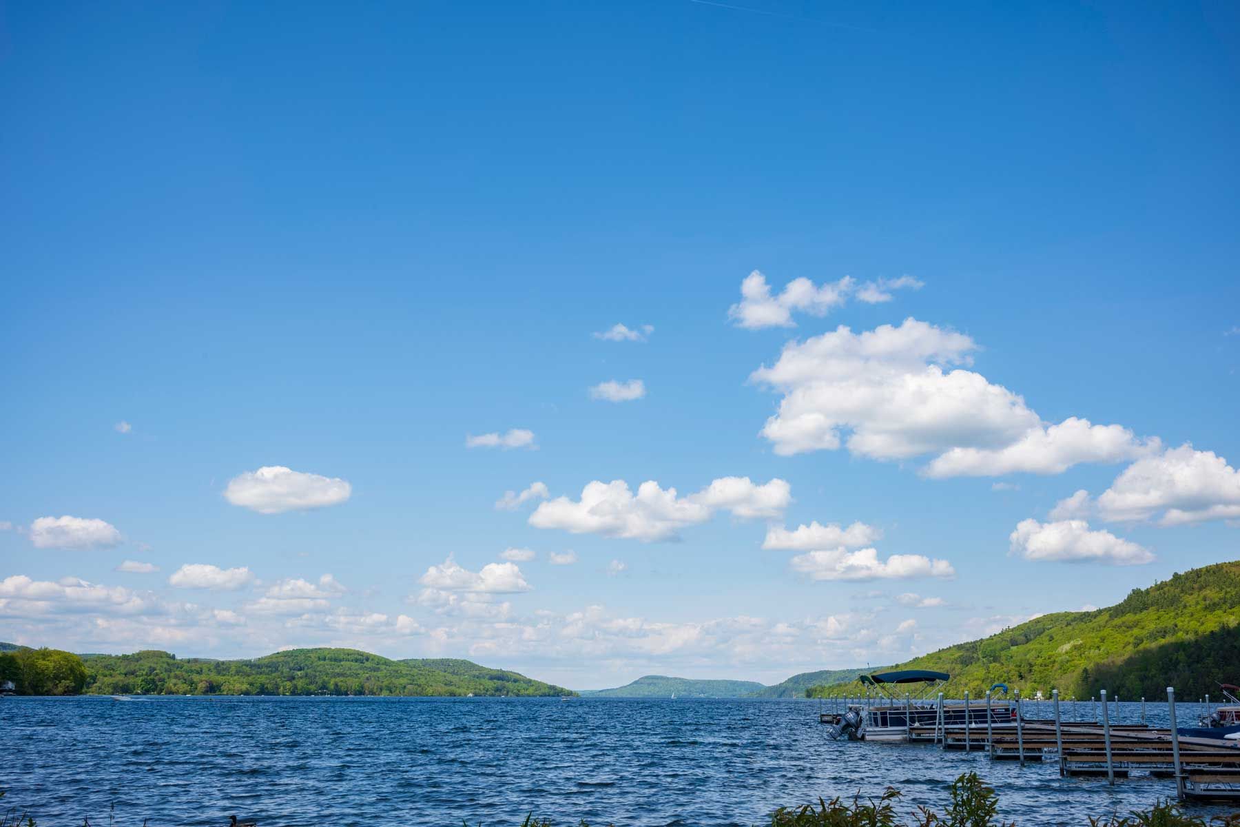 Das blaue Wasser des Otsego Lake in Cooperstown, New York, an einem sonnigen Sommertag mit Kumuluswolken am Himmel, fotografiert in der Nähe eines Docks.